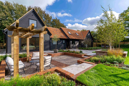 Barn conversion with black timber cladding, red tiled roof, and landscaped courtyard featuring pergola seating area