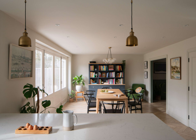 Dining room with wooden table, black chairs, wall-mounted bookshelves, and large windows allowing natural light.