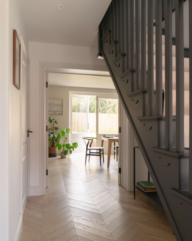 Hallway with dark staircase and herringbone flooring, leading to dining area with large windows.