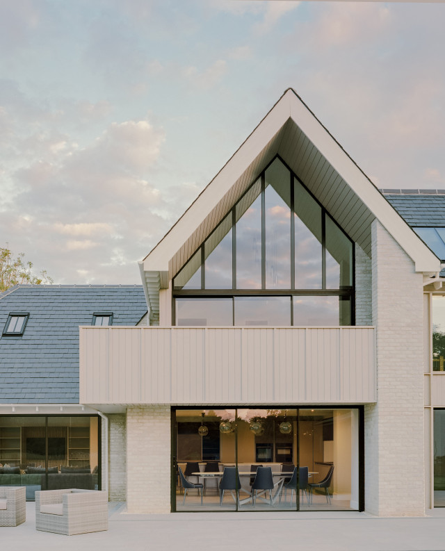 Contemporary house facade featuring a tall glazed gable, first-floor balcony with vertical cladding, and large ground-floor sliding doors beneath a pitched roof
