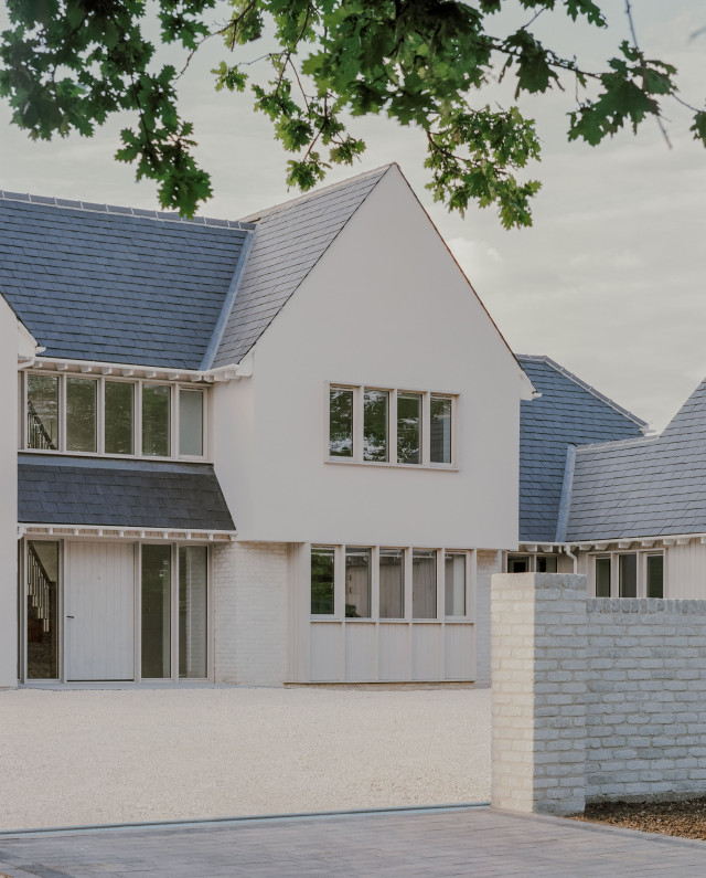Modern house with pitched roofs, pale render, brick detailing, and a row of slender windows overlooking a gravelled front area.