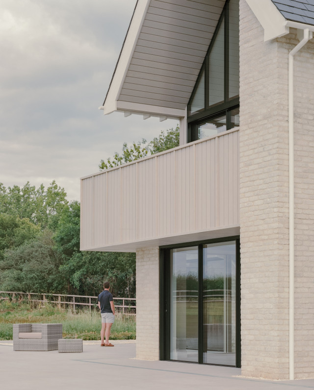 Modern house elevation with light brickwork, full-height glazing, and a projecting first-floor balcony featuring vertical timber cladding.