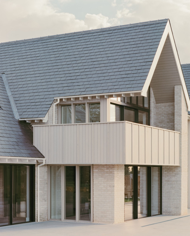 Contemporary house with light brick walls, slate-tiled gable roof, and a cantilevered first-floor balcony clad in vertical pale timber panels above large sliding glass doors