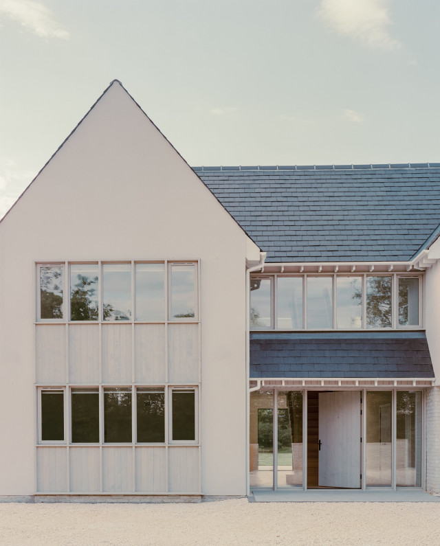 Contemporary house with a tall rendered gable, large vertically aligned windows with timber surrounds, and an adjoining wing featuring a slate roof and a recessed glazed entrance